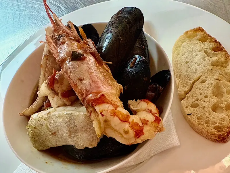A bowl of seafood stew featuring shrimp, mussels, and fish, served with rustic bread. Experience authentic Italian cuisine Ribble Valley style—perfect for an Italian wine pairing dinner. Presented on a white plate with a napkin under the bowl.