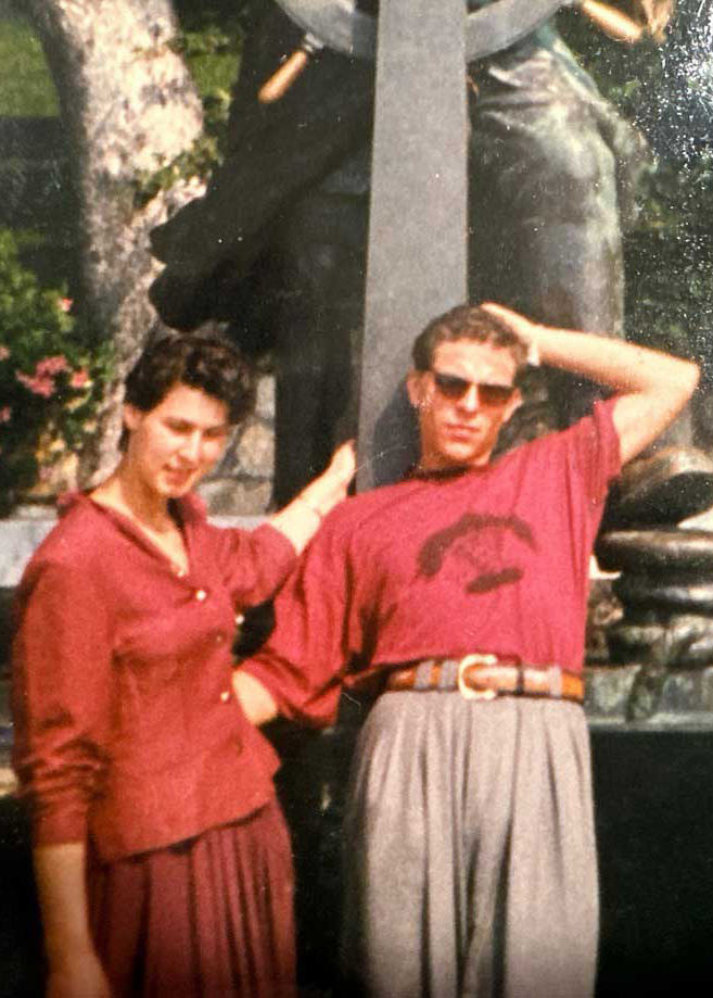 A woman and man pose together outdoors in front of a statue, celebrating La Locanda 22nd anniversary. She wears a maroon blouse and skirt; he sports sunglasses with a maroon shirt tucked into high-waisted, belted gray trousers.