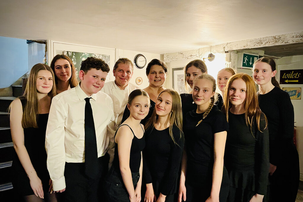 A group of young people and two adults from Stage Door Youth Theatre pose together indoors. Most are wearing black clothes, some are smiling, and they stand closely in a warmly lit room at the La Locanda charity event.