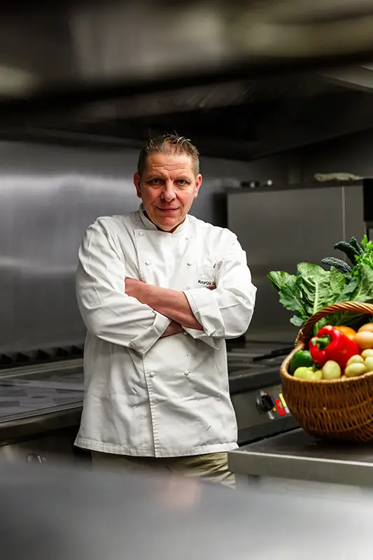 A chef in a white uniform stands with arms crossed in a commercial kitchen, next to a basket of fresh vegetables—peppers, tomatoes, and leafy greens—at La Locanda Maurizio Bocchi, home of the Maurizio Bocchi Tourism Superstar.