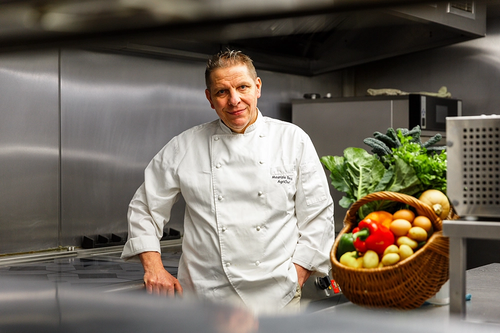 Chef Maurizio Bocchi, nominee for the VisitEngland Tourism Superstar Awards 2026 stands in his modern commercial kitchen, smiling next to a basket filled with fresh vegetables including peppers, tomatoes, potatoes, and leafy greens.