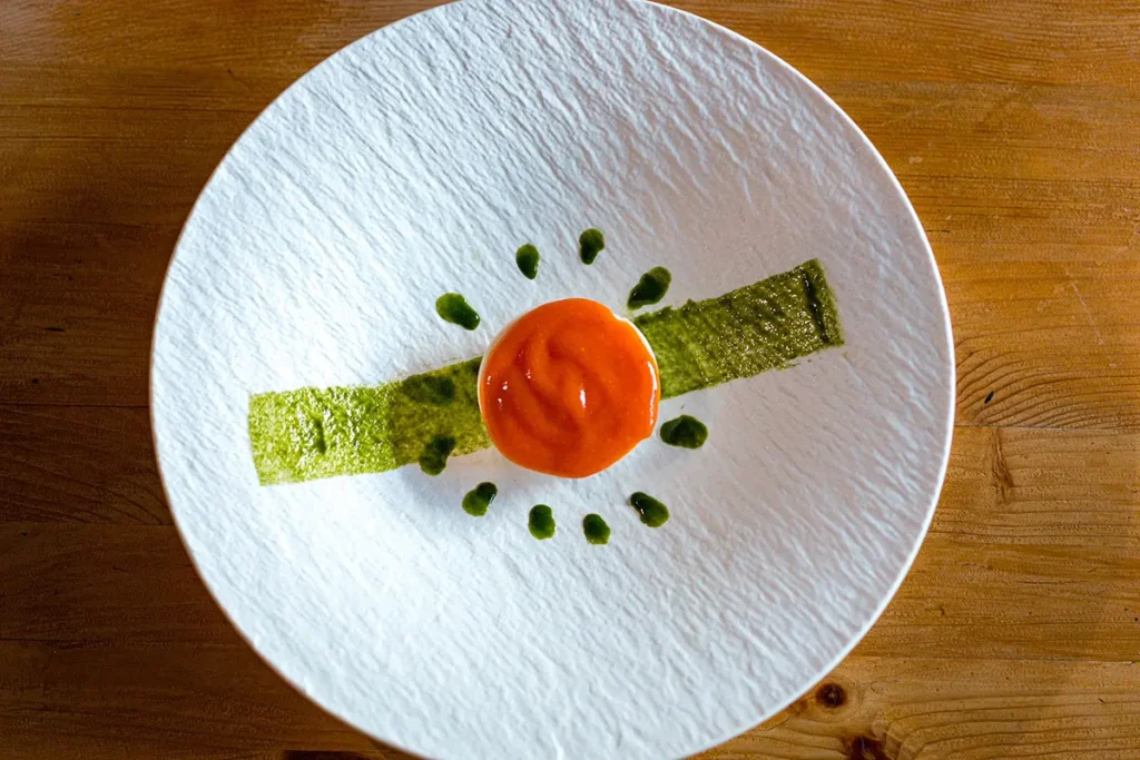 A white textured plate with a green rectangular strip, Burrata Semifreddo with tomato coulis in the centre, and small green dots arranged in a semi-circle, all on a wooden table.